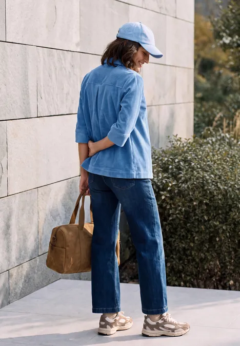 Femme en blazer et casquette bleu clair, jean et baskets, de profil contre un mur, sac marron dans le dos.