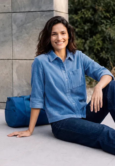 Une femme souriante assise devant un mur, en chemise en jean, une sacoche bleue posée à côté d'elle.