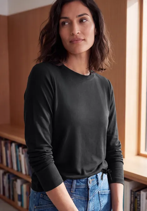 Eine Frau mit schwarzem Langarmshirt und Jeans steht lässig vor einem Bücherregal.