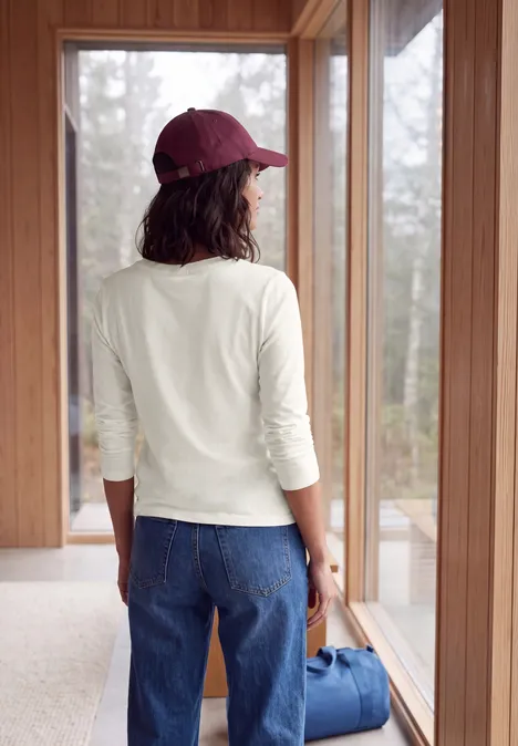 Eine Frau steht mit Jeans, weißem Shirt und roter Kappe vor einem Fenster und hält eine blaue Tasche.