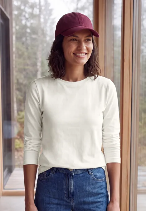 Frau in weißem Langarmshirt, Jeans und roter Mütze steht lächelnd vor großen Fenstern mit Waldblick.