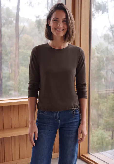 Langarm Shirt mit Tunnelzug am Saum truffle brown Frau in lässiger Jeans und Pullover steht vor großen Fenstern mit ruhiger Waldkulisse.