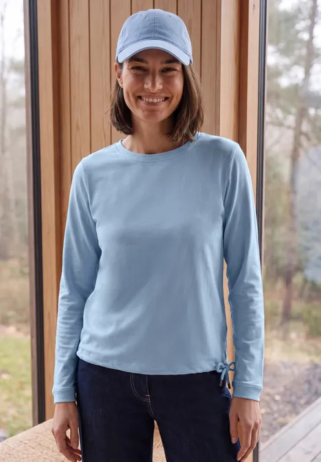 Een lachende vrouw draagt een lichtblauw shirt met lange mouwen, jeans en pet, staand voor een houten raam.