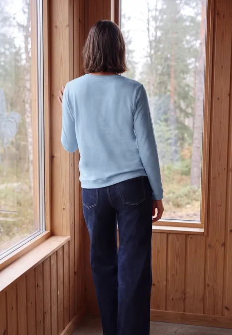 Een persoon staat in een houten kamer en kijkt door grote ramen naar het bos.