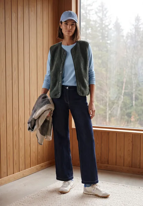 Vrouw in casual outfit met bodywarmer, pet, jas in de hand, wijde jeans en sneakers in een houten kamer voor het bos.