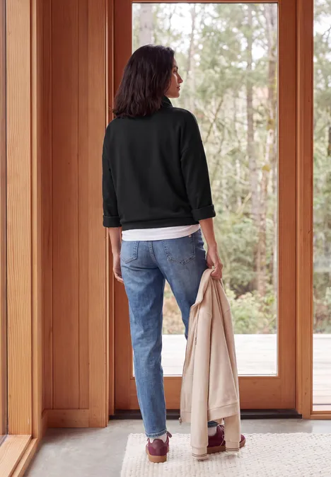 Eine Person von hinten in Jeans und Jacke hält einen Mantel und blickt durch eine Glastür.