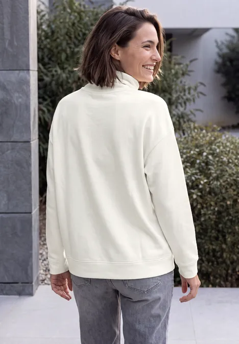Vrouw in crème fleecevest en grijze jeans, glimlachend van opzij kijkend buiten.