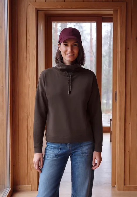 Een vrouw met paarse pet, donker sweatshirt en blauwe jeans lacht in een houten deuropening.
