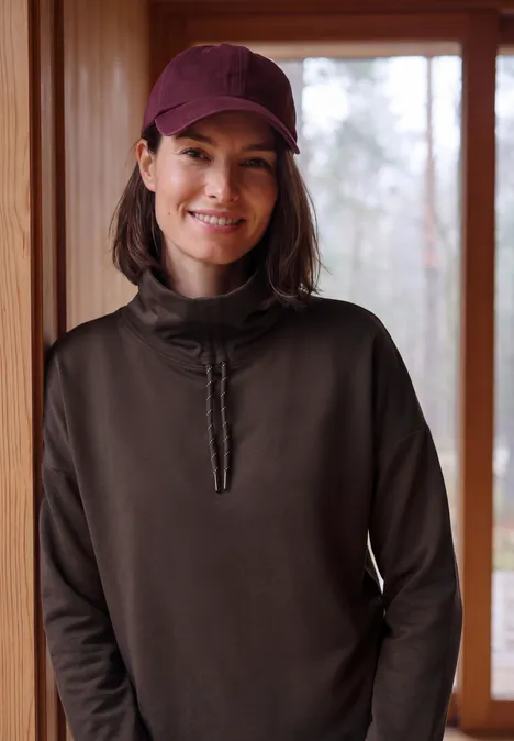 Vrouw met rode pet en donker trui lacht voor een raam in een houten interieur.