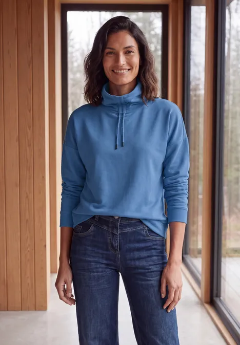 Een vrouw in een blauw trui en spijkerbroek staat voor grote ramen in een houten ruimte en lacht.