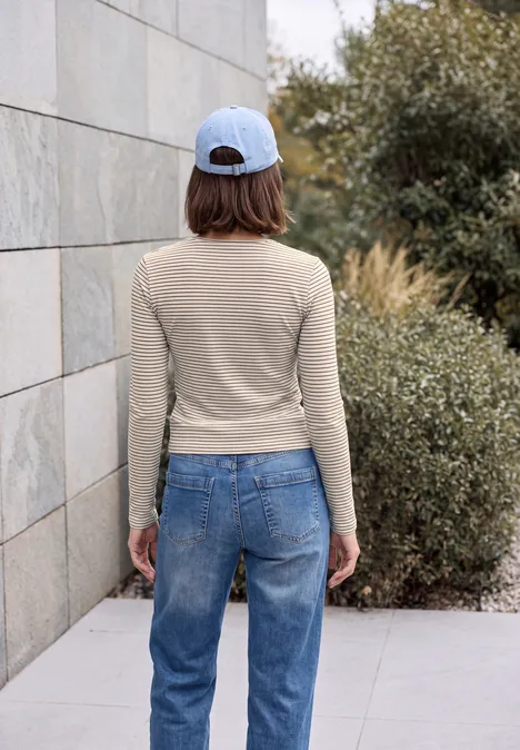 Frau steht mit dem Rücken zur Kamera, trägt Jeans, Beige-Langarmshirt und Baseballcap, vor einer Mauer und Büschen.