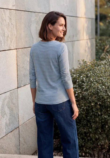Frau in grauem Dreiviertelshirt und blauer Jeans lehnt an einer Steinwand und schaut seitlich.