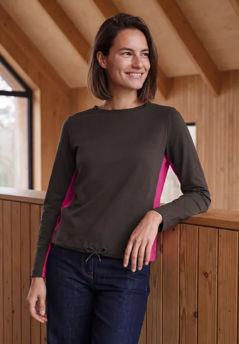Une femme souriante en haut manches longues avec inserts roses appuyée sur une rambarde dans un chalet en bois.