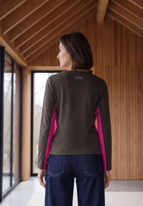 Femme dans une pièce en bois, portant un pull olive aux côtés roses et un jean.