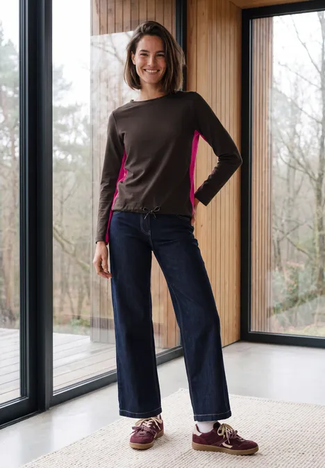 Une femme souriante en sweat moderne et jean large, debout devant de grandes fenêtres dans une pièce lumineuse.