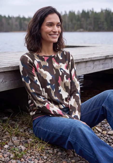 T-shirt à manches longues, col bateau et imprimé pink flash Eine Frau sitzt lächelnd am Holzsteg an einem See, trägt Camouflage-Pullover und Jeans.