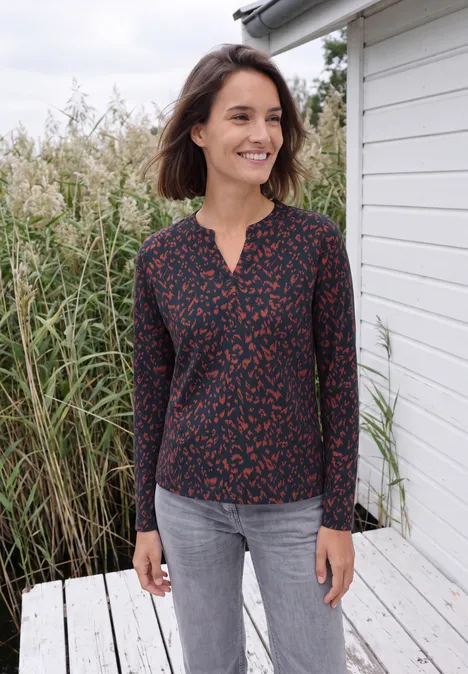 Shirt mit Split Neck im Tunika-Look urban dark blue Frau in dunkelblauer, rot bedruckter Bluse und grauen Hosen steht lächelnd vor einem weißen Haus und Schilf.