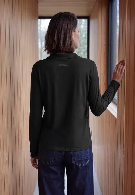 Langarm Shirt mit Hemdkragen Black Person von hinten in schwarzem Langarmshirt und Jeans, steht im hellen Holzflur am Fenster.