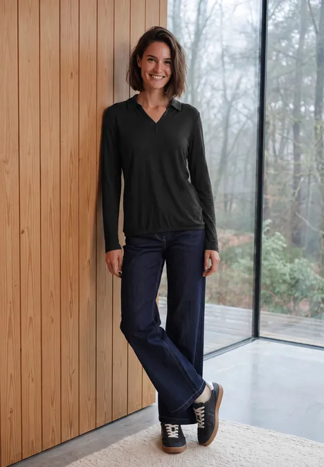 Langarm Shirt mit Hemdkragen Black Eine Frau lehnt lächelnd in Jeans und schwarzem Langarmshirt an einer Holzwand vor großen Fenstern.