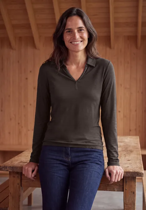 Femme souriante appuyée sur une table en bois devant un mur en lambris, porte un haut à manches longues et un jean.