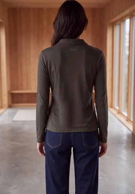 Femme de dos portant un t-shirt vert à manches longues et un jean bleu dans une pièce en bois lumineuse.