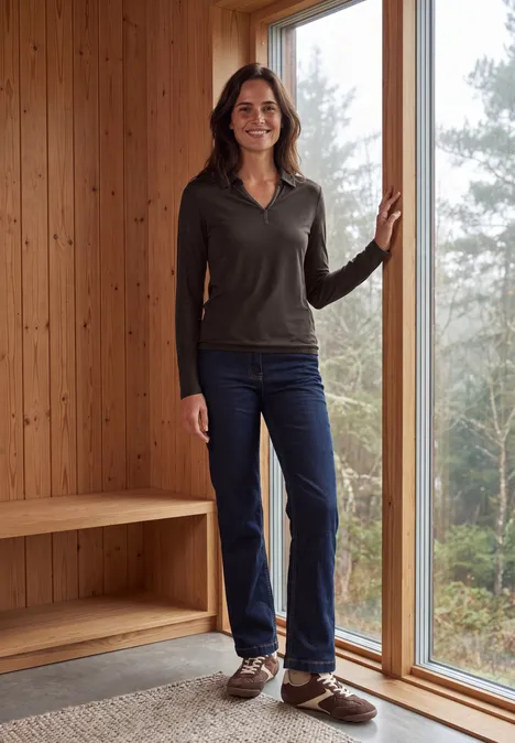 Femme en tenue décontractée (pull, jean, chaussures marron) debout près d'une baie vitrée dans un intérieur bois.