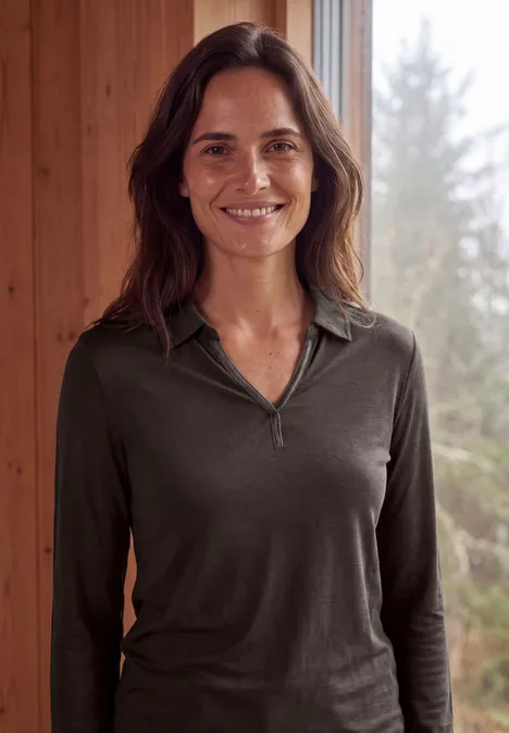 Une femme souriante aux cheveux bruns porte un haut vert foncé devant une fenêtre offrant une vue sur la forêt.