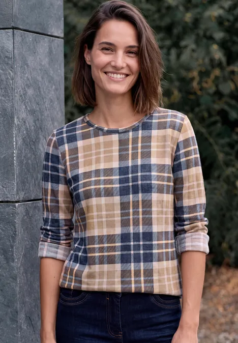 Eine lächelnde Frau mit kariertem Pullover lehnt im Freien an einer Wand.