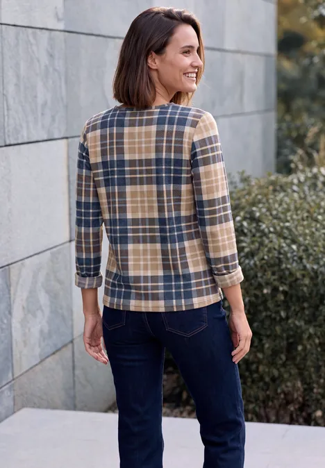 Eine Frau mit braunen Haaren in karierter Bluse und dunkler Jeans steht lächelnd vor Mauer und Büschen.