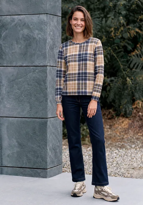 Eine Frau in kariertem Pullover und Jeans steht lächelnd vor einer grauen Wand und Grünpflanzen.