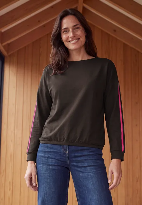 Shirt mit Boat Neck und Tapedetails truffle brown Eine lächelnde Frau in Jeans und langärmeligem Oberteil steht vor einer Holzwand.
