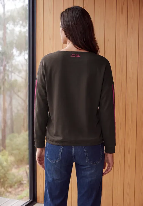 Shirt mit Boat Neck und Tapedetails truffle brown Frau von hinten in dunklem Sweatshirt und Jeans, steht vor Holzwand und Blick auf bewaldete Umgebung.