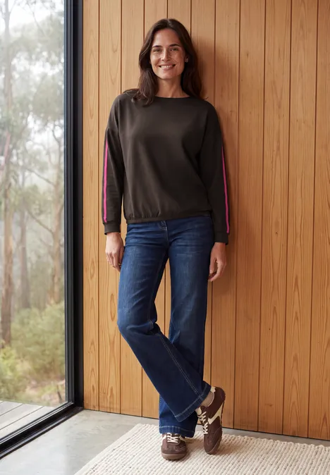 Shirt mit Boat Neck und Tapedetails truffle brown Eine lächelnde Frau in Jeans und Pullover steht in einem hellen Holzraum neben einer bodentiefen Fensterscheibe.