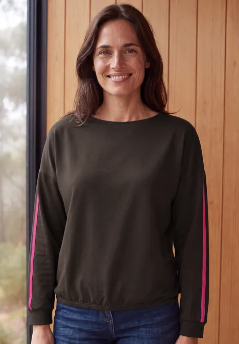 Shirt mit Boat Neck und Tapedetails truffle brown Eine lächelnde Frau in grünem Sweatshirt und Jeans vor einer Holzwand.