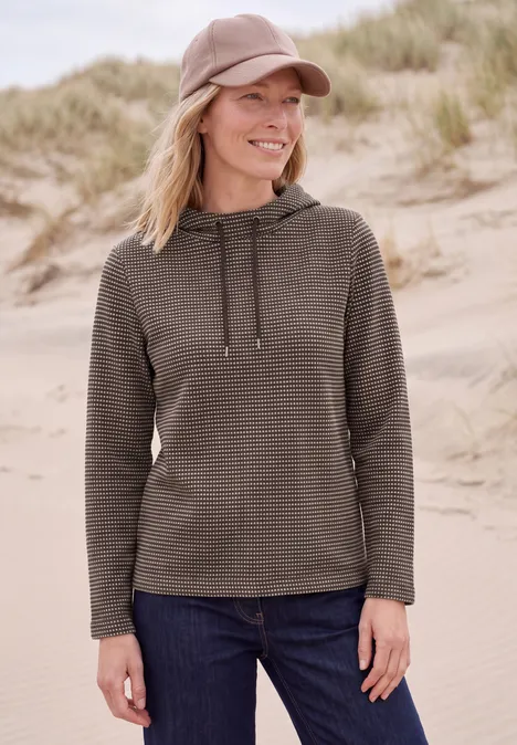 T-shirt à manches longues et col cheminée truffle brown Eine lächelnde Frau in grüner Kapuzenjacke, Jeans und Cap steht vor sandigen Dünen am Strand.