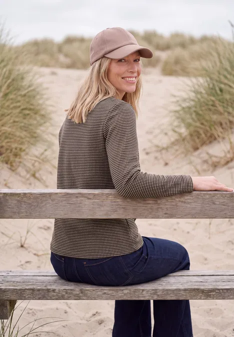 T-shirt à manches longues et col cheminée truffle brown Frau in Pullover und Jeans sitzt auf Holzbank im Dünenstrand, blickt lächelnd zur Kamera.