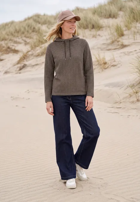T-shirt à manches longues et col cheminée truffle brown Frau mit Kapuzenpullover und weitem Jeans auf Dünenstrand, schaut lächelnd zur Seite.