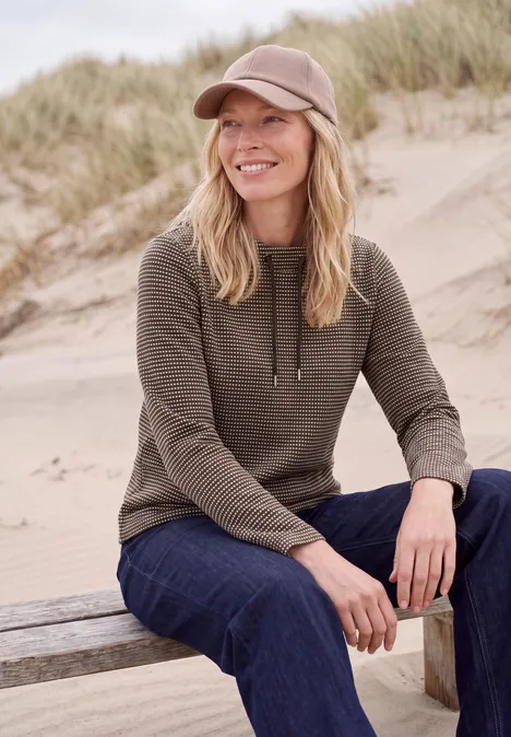 T-shirt à manches longues et col cheminée truffle brown Eine lächelnde Frau in Baseballkappe und Strickpullover sitzt entspannt auf einer Düne.