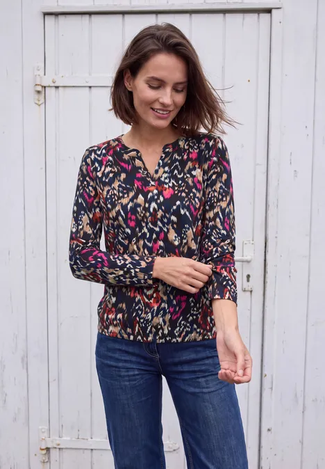 Shirt mit Split Neck im Tunika-Look truffle brown Frau in Blumenshirt und Jeans vor weißer Holztür, lächelnd und Ärmel zurechtrückend.