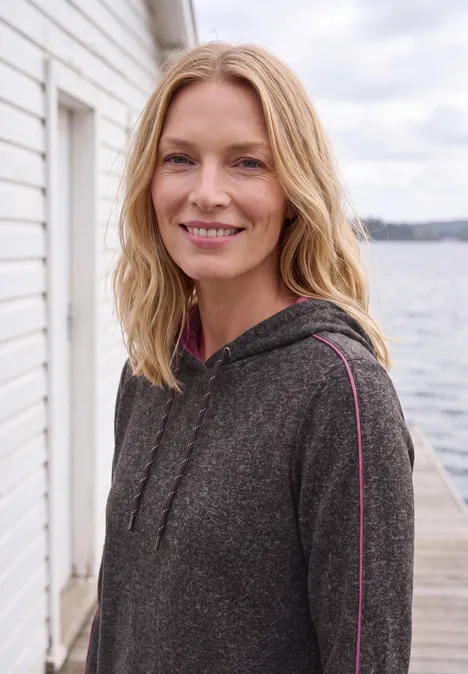 T-shirt à manches longues avec capuche et passepoil truffle brown melange Eine lächelnde Frau mit blonden Haaren in einem grauen Hoodie vor einem Bootshaus und Gewässerkulisse.