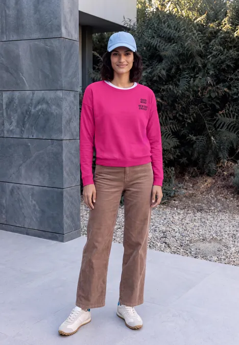 Vrouw in roze sweater, bruine broek, witte sneakers en pet voor een modern huis met struiken.