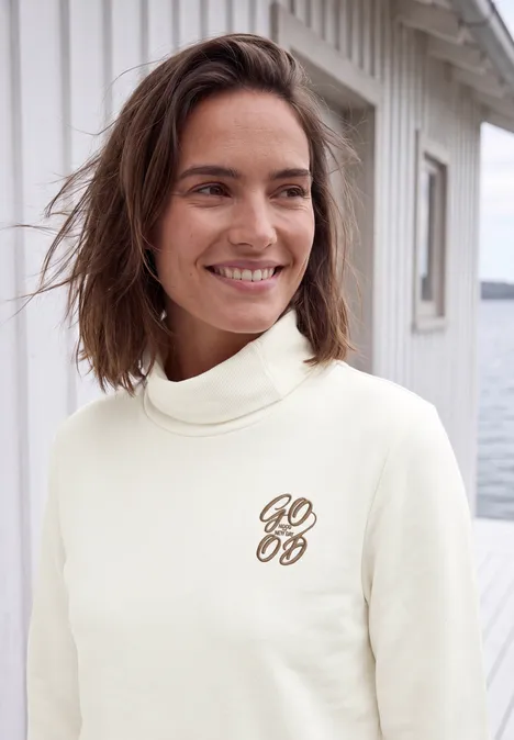 Koszulka z długim rękawem, golfem i nadrukiem vanilla white Kobieta w jasnym golfie z eleganckim logo, uśmiechnięta przed białym drewnianym domkiem nad wodą.