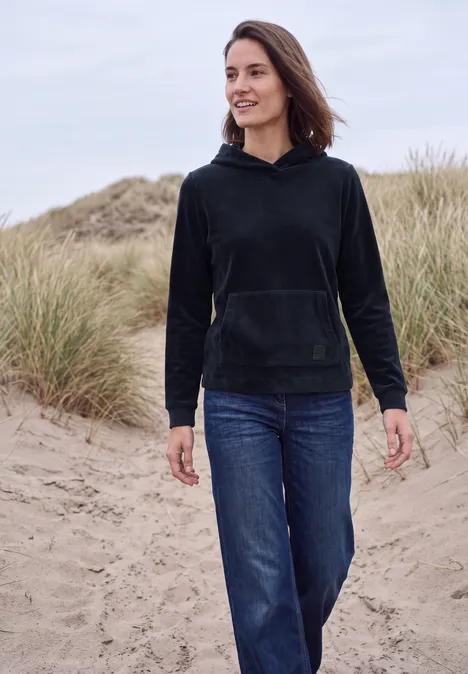 Langarm Shirt aus Cord mit Kapuze urban dark blue Eine Frau in schwarzer Kapuzenjacke und Jeans spaziert am Strand zwischen Dünen.