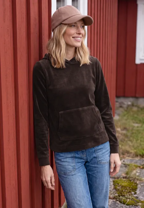 Langarm Shirt aus Cord mit Kapuze truffle brown Eine Frau in dunkelbraunem Hoodie, Jeans und Cap lehnt lächelnd an einer roten Holzwand.
