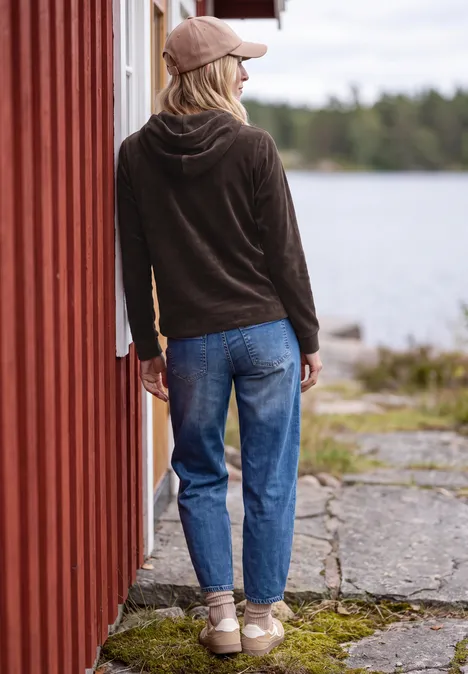 Langarm Shirt aus Cord mit Kapuze truffle brown Person lehnt mit dem Rücken an einem roten Holzhaus am See, trägt Jeans, Kapuzenpullover und Cap.