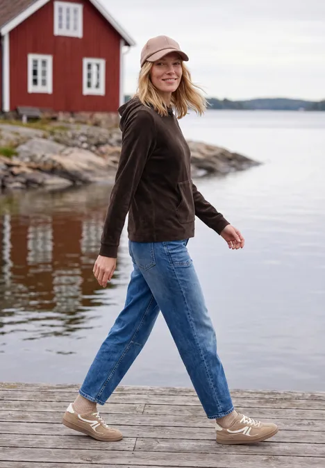 Langarm Shirt aus Cord mit Kapuze truffle brown Eine Frau mit Jeans und Jacke spaziert auf einem Steg am See vor einem roten Haus und lächelt.