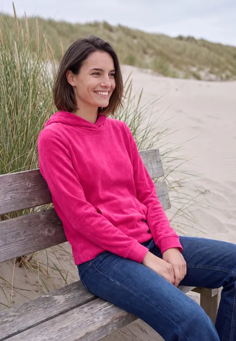 T-shirt à manches longues en velours côtelé avec capuche pink flash Eine Frau in pinkem Hoodie und Jeans sitzt lächelnd auf einer Holzbank am Strand vor Dünen.