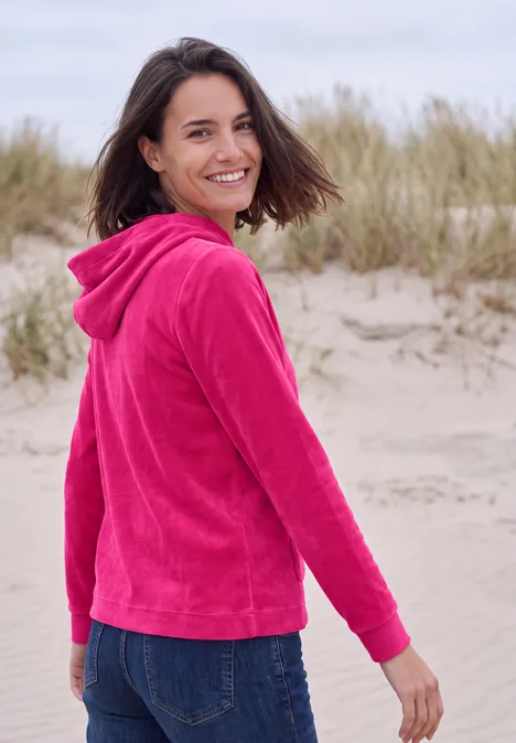 T-shirt à manches longues en velours côtelé avec capuche pink flash Eine Frau in pinkem Hoodie und Jeans steht auf Sanddünen am Strand und lächelt zurück.