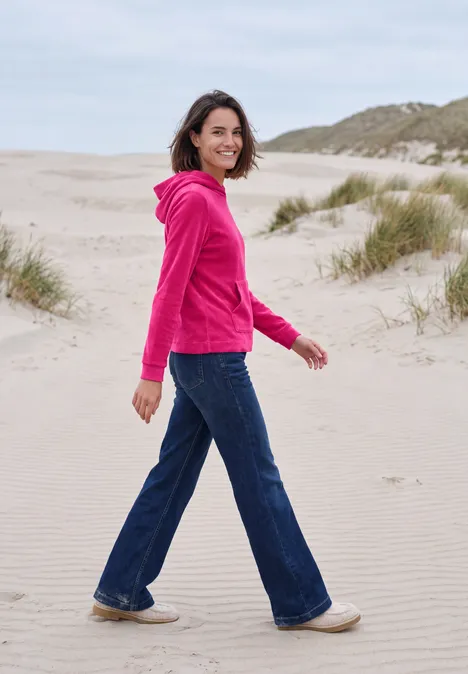 T-shirt à manches longues en velours côtelé avec capuche pink flash Eine Frau in pinkem Kapuzenpullover und Jeans läuft am Strand.