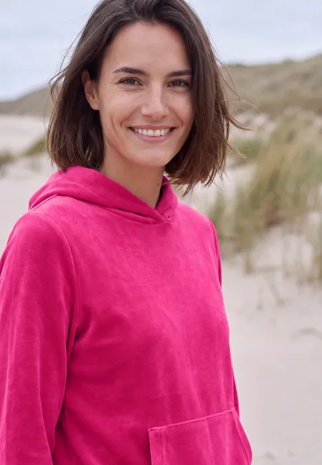 T-shirt à manches longues en velours côtelé avec capuche pink flash eine lächelnde Frau in pinkem Hoodie steht am Strand, im Hintergrund Dünen zu sehen.